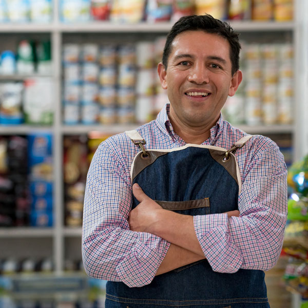A small business owner standing inside his grocery store with his arms across his chest and a big smile on his face. Behind him are shelves filled with groceries.